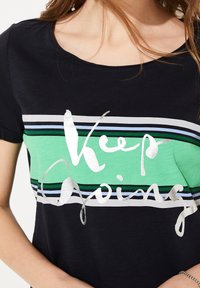 Sortærmet t-shirt i sort med grønne og blå vandrette striber. Har sølvtrykt tekst "Keep Going" på brystet.