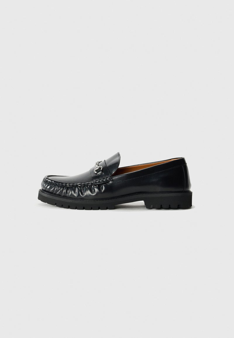 Mocasín de cuero negro con punta redondeada, costuras decorativas y un acento metálico en la parte delantera; suela de goma con patrón de tracción.