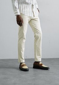 Hvidstribet skjorte med knapper, cremede slim-fit bukser og tofarvede loafers med vævet front og brune accenter, sat op imod en grå baggrund.