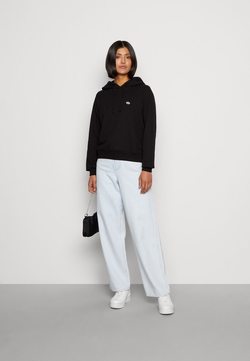 Schwarzer Kapuzenpullover mit Logo, kombiniert mit hellblauen weit geschnittenen Jeans und weißen Sneakers. Die Person hält eine kleine schwarze Tasche.