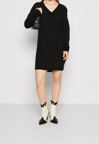 Abito lavorato a maglia nero con scollo a V e maniche lunghe, abbinato a stivali da cowboy crema e neri con punta affilata e tacco alto.