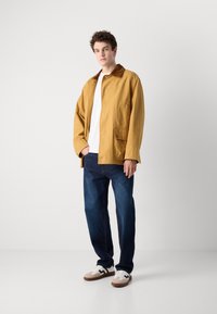 Giacca beige di cotone con colletto marrone, tasche anteriori, camicia bianca, jeans blu e sneakers marroni e bianche, in piedi contro uno sfondo chiaro.