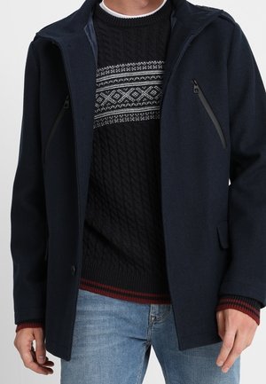 Homme portant une veste bleu marine par-dessus un pull en maille noir avec un motif blanc, assorti à un jean bleu clair.