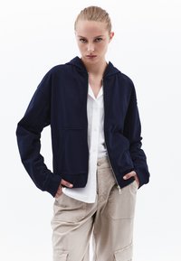Felpa con zip di colore blu navy, dotata di una tasca frontale, polsini a coste e una vestibilità casual, indossata sopra una camicia bianca con bottoni e pantaloni cargo beige.