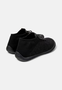 Chaussures de sport noires en tissu texturé, avec un design bas et des lacets, inclinées pour montrer le talon et le profil latéral.