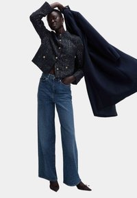 Giacca crop nera con texture e bottoni dorati, abbinata a jeans in denim blu a gamba larga. Cappotto scuro drappeggiato su un braccio.