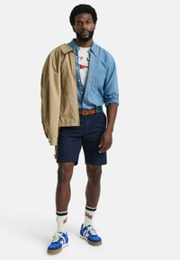 Blå denimskjorte over en hvit grafisk t-skjorte, beige lett jakke, marineblå shorts og blå sneakers med hvite detaljer og stripete sokker.