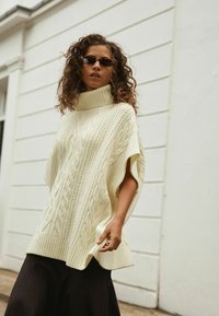 Cremfarbener gestrickter Poncho mit hohem Kragen, der Kabelmuster und kurze Ärmel aufweist, über einem schwarzen Rock. Das Modell trägt eine Sonnenbrille.