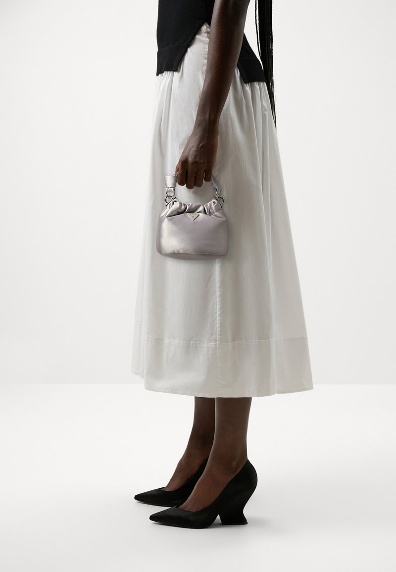 Silvery satin handbag with gathered top, held by a hand. Worn with a flowing white skirt and black wedge heels, showcasing a minimalist design.