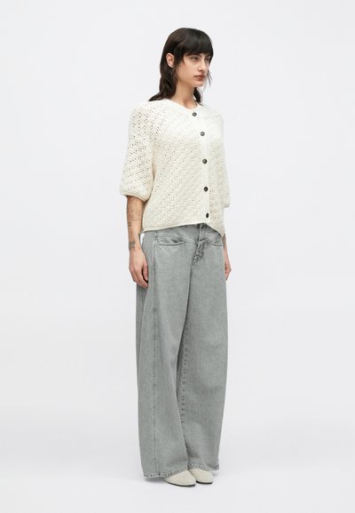 Πλεκτή, εκρού ζακέτα με κοντά, φουσκωτά μανίκια και κουμπιά. Συνδυάζεται με ανοιχτό γκρι, φαρδιές τζιν παντελόνες και μπεζ παπούτσια.