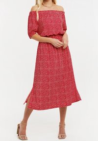 Robe rouge à épaules dénudées avec motif à pois blancs, manches courtes bouffantes, taille élastique, et fentes latérales hautes jusqu'à la cuisse. Texture de tissu lisse.