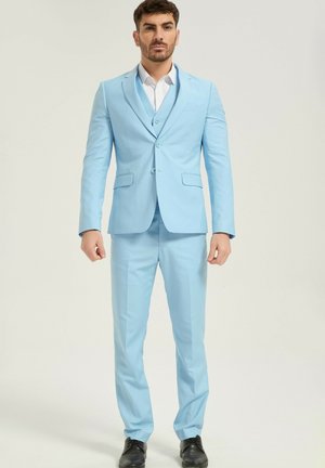 Homme debout, face à l'avant, portant un costume trois pièces bleu clair avec des chaussures noires et une chemise blanche sur un fond uni.