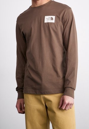 Mann trägt ein braunes Langarmshirt von The North Face mit Logo-Aufnäher auf der Brust und senfgelbe Hose vor einfachem Hintergrund.