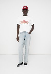Junger Mann mit rotem Hut, weißem T-Shirt mit Grafik, hellblauen Jeans und schwarzen Sneakers, der vor einem schlichten weißen Hintergrund steht.