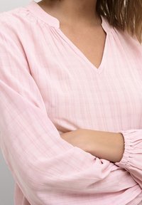 Blouse rose clair avec un col en V et des détails froncés, dotée de longues manches avec des poignets élastiques et une texture à carreaux.