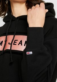 Persona con sudadera negra con el logo rosa de "TOMMY JEANS", sosteniedo la manga cerca del cuello, pequeña bandera de Tommy Hilfiger visible en el puño de la muñeca.