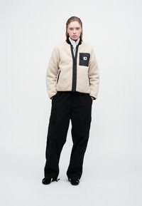 Beige fleece-takki mustilla yksityiskohdilla, edessä vetoketju ja läppätasku. Yhdistetty löysiin mustiin cargo-housuihin, joissa on sivutaskut.