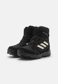 Botas de senderismo negras con rayas blancas, que cuentan con una parte superior texturizada, cierre de cordones y una correa de velcro para mayor soporte. Suela de goma para tracción.