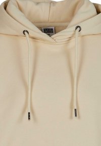 Sweat à capuche beige avec un cordon de serrage à embout métallique, texture douce, et une étiquette "Urban Classics XS" à l'intérieur du col.
