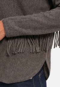 Siv krožen pulover z dolgimi resicami na rokavih. Mehka tekstura in rahlo ukrivljen rob, v kombinaciji s temnimi jeans hlačami.