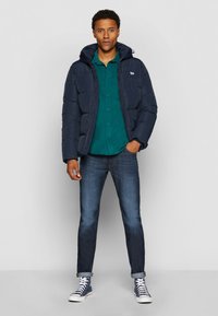 Doudoune marine avec capuche, chemise verte à boutons en dessous, jeans foncés et baskets blanches. Tenue décontractée en couches.