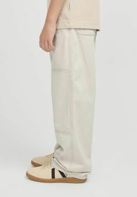 Bež laiad püksid kontrastõmbluste, küljetaskute ja pehme tekstuuriga, mida on kombineeritud heledate tossudega mustade triipudega.