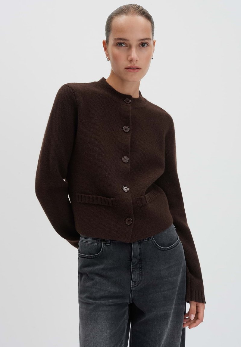 Woman wearing a dark brown buttoned cardigan with front pockets and faded black jeans, standing against a plain light background.