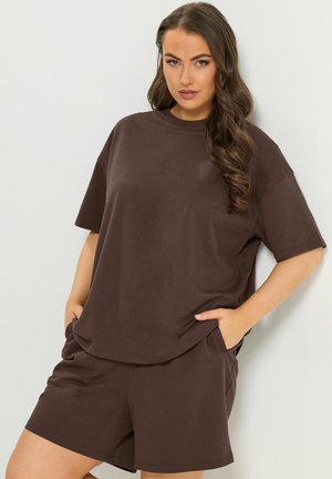 Kobieta z długimi falującymi włosami, ubrana w luźny ciemnobrązowy T-shirt i pasujące szorty, z rękami w kieszeniach, stojąca na tle białej ściany.