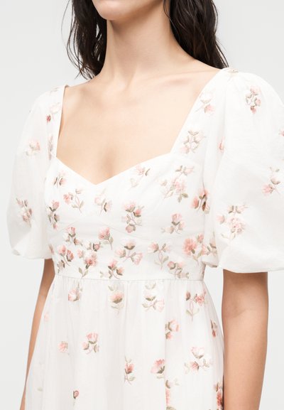 Γυναίκα φοράει λευκό φόρεμα με φουσκωτά μανίκια που διαθέτουν λεπτή ροζ και πράσινη κεντητή λουλουδένια διακόσμηση, με λαιμόκοψη σε απαλό τετράγωνο στυλ.