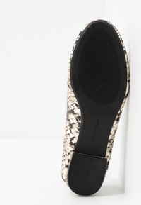 Semelle d'une chaussure pour femmes avec un talon et un avant en caoutchouc noir texturé, tige en motif serpent beige et noir, placée verticalement contre un mur blanc.