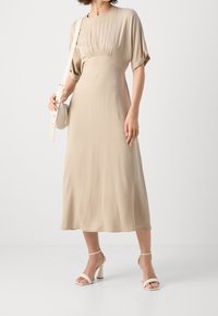 Béžové midi šaty se shromážděním v pase, krátkými rukávy a hladkou texturou; doplněné o sandály na vysokém podpatku se zavazováním a kulatou kabelkou přes rameno.
