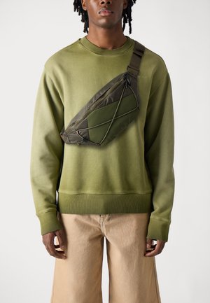 Felpa verde oliva, strutturata, con polsini a costine, abbinata a pantaloni corti beige chiaro. Sulla spalla c'è una borsa a tracolla verde oliva scuro con un motivo geometrico.
