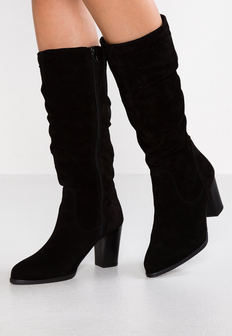 Bottes mi-mollet en daim noir avec talons blocs et fermetures éclair latérales, portées sur des jambes nues, sur un fond blanc uni.