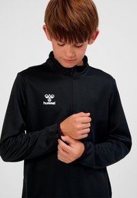 Junge mit braunen Haaren, der ein schwarzes Hummel-Sportoberteil mit Reißverschluss trägt und den Ärmelbündchen vor einem schlichten weißen Hintergrund anpasst.