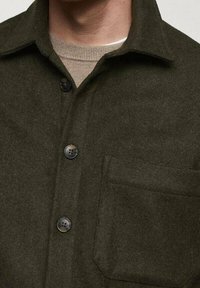 Mørkegrøn uldskjorte med spidst revers, sorte knapper og en lomme foran. Blød tekstur, skræddersyet pasform og minimalistisk design.