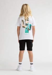T-shirt blanc avec un motif graphique coloré au dos, accompagné d'un short noir et de baskets blanches avec des accents noirs.
