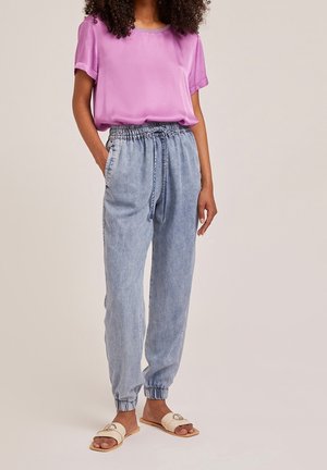 Kvinde iført en løs lyserød kortærmet bluse, lyseblå denim joggers med elastisk talje og beige åbne flade sandaler.