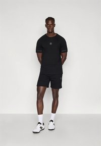 T-shirt e calções atléticos pretos com logótipo, feitos de tecido leve. Usados com sapatos brancos com detalhes em preto e dourado.