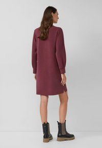 Burgunderfarbenes Sweatshirtkleid mit langen Ärmeln, rundem Ausschnitt, seitlichen Schlitzen und weicher Textur, kombiniert mit schwarzen Knöchelstiefeln mit dicker Sohle.