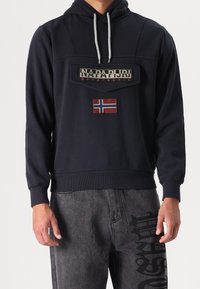 Mężczyzna w czarnej bluzie Napapijri z naszywką w norweski flagę oraz ciemnoszarych dżinsach z czarnym graficznym napisem na udzie.