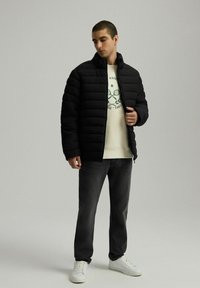 Czarna kurtka puchowa z poziomymi przeszyciami, wysoki kołnierz, w połączeniu z kremowym swetrem i ciemnymi dżinsami. Model nosi białe trampki.