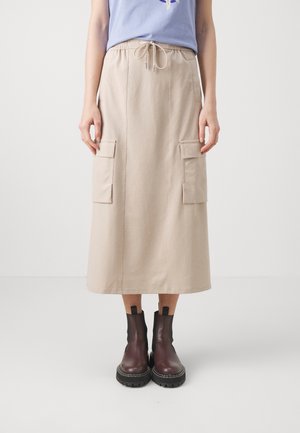 Kobieta ubrana w beżową midi spódnicę z przednimi kieszeniami i ściągaczem w talii, niebieską bluzkę oraz brązowe masywne skórzane botki, stojąca na białej podłodze.