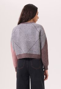 Pull en tricot gris et rose au design court, avec des motifs géométriques, un ourlet côtelé et des manches longues. Porté avec un jean foncé.