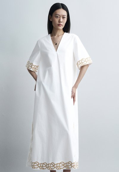 Femme aux cheveux noirs raides portant une robe blanche ample avec une broderie circulaire beige sur les manches et l'ourlet, debout devant un fond uni.