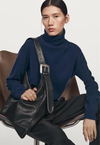 Zwarte leren schoudertas met een grote opening, zilveren hardware en een brede verstelbare schouderband. Model draagt een donkerblauwe coltrui en broek.