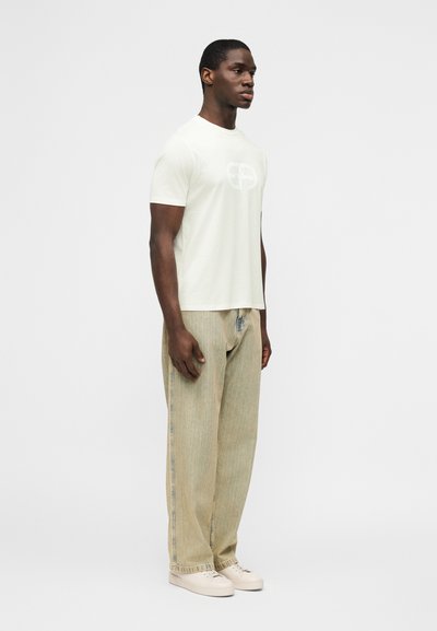 Άνδρας που στέκεται φορώντας ένα λευκό t-shirt με κοντά μανίκια και λογότυπο, μπεζ φαρδιά τζιν και εκρού αθλητικά παπούτσια μπροστά σε απλό φόντο.