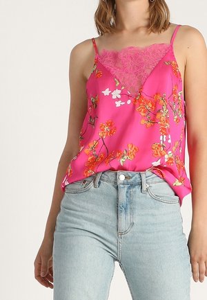 Kobieta w jasnoróżowej kwiecistej koszulce z koronkową lamówką, zestawiona z jasnoniebieskimi jeansami z wysokim stanem, stoi na tle jednolitego tła.