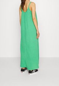 Zielona, bezrękawnikowa sukienka maxi z teksturowanej tkaniny, o luźnym kroju i marszczonym detalu z tyłu, zestawiona z czarnymi tenisówkami.