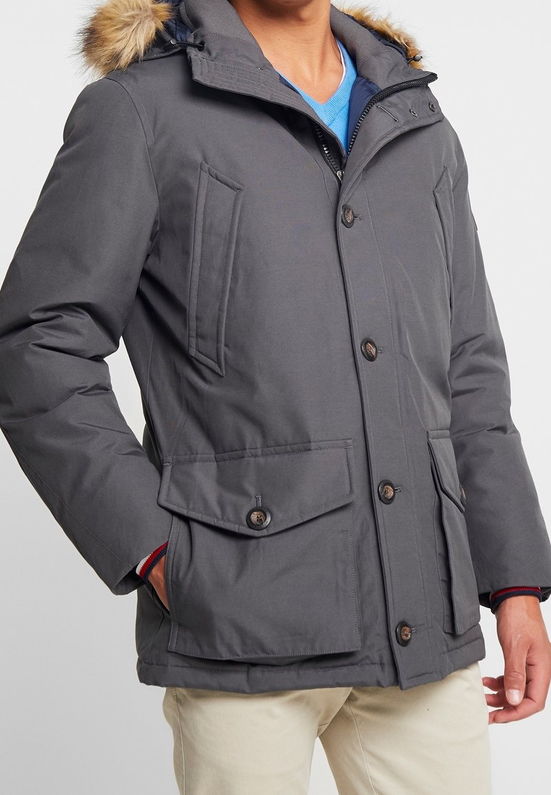 Homme portant une veste d'hiver grise avec capuche bordée de fourrure, devant boutonné, poches à rabat, chemise bleue et pantalon beige, debout avec une main dans la poche.