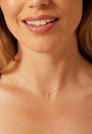 Collier en chaîne dorée avec un petit pendentif stylisé en forme de lettre "J". Design délicat, texture lisse, et maillons fins reposant sur une clavicule nue.